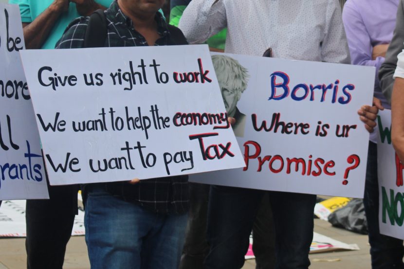 Two people holding a signs at the Solidarity Rally For The Regularisation Of Undocumented Migrants held in July 2021 outside 10 Downing Street. The sign on the rights says: 'Give us right to work. We want to help the Economy. We want to pay Tax.' The sign on the right includes a hand drawn picture of UK Prime Minister, Boris Johnson, and says: 'Boris where is ur Promise?